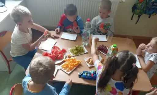 Dzieci siedzą w kółku przy stole zastawionym owocami i innymi przekąskami.